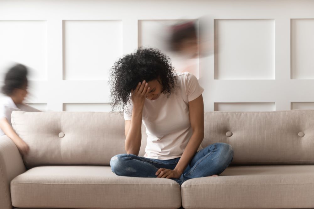 A fatigued woman sitting on a couch, her head in her hand, showing signs of anemia while children move in the background