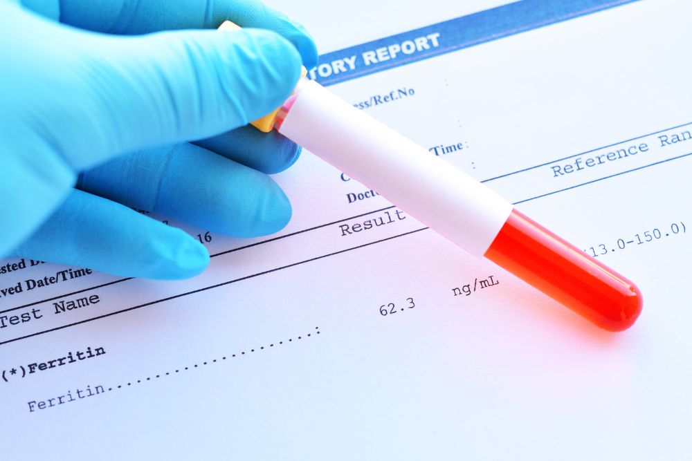 A gloved hand holding a blood sample vial placed on top of a lab report showing ferritin levels, indicating iron-related health testing.