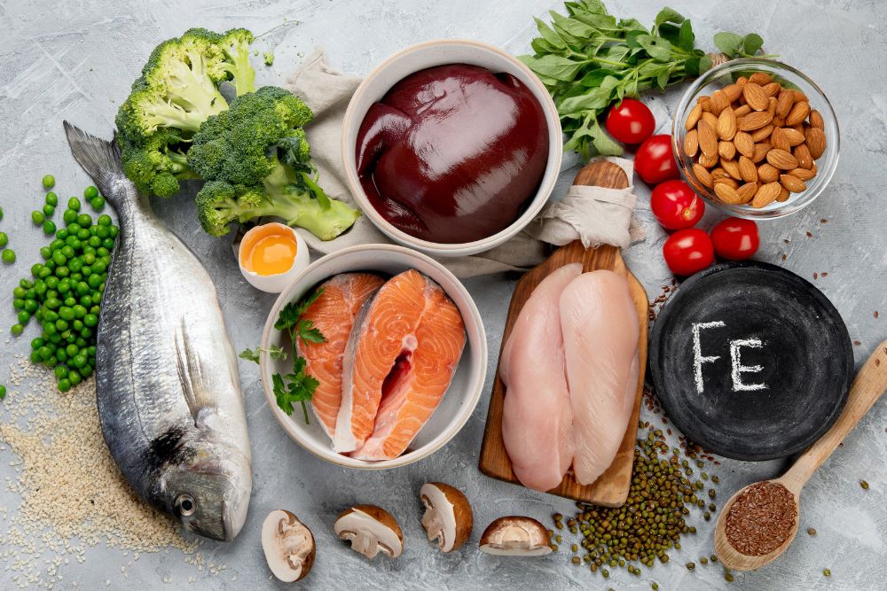 A variety of iron-rich foods, including fish, liver, chicken, salmon, broccoli, almonds, and leafy greens, arranged on a table to promote dietary sources of iron.