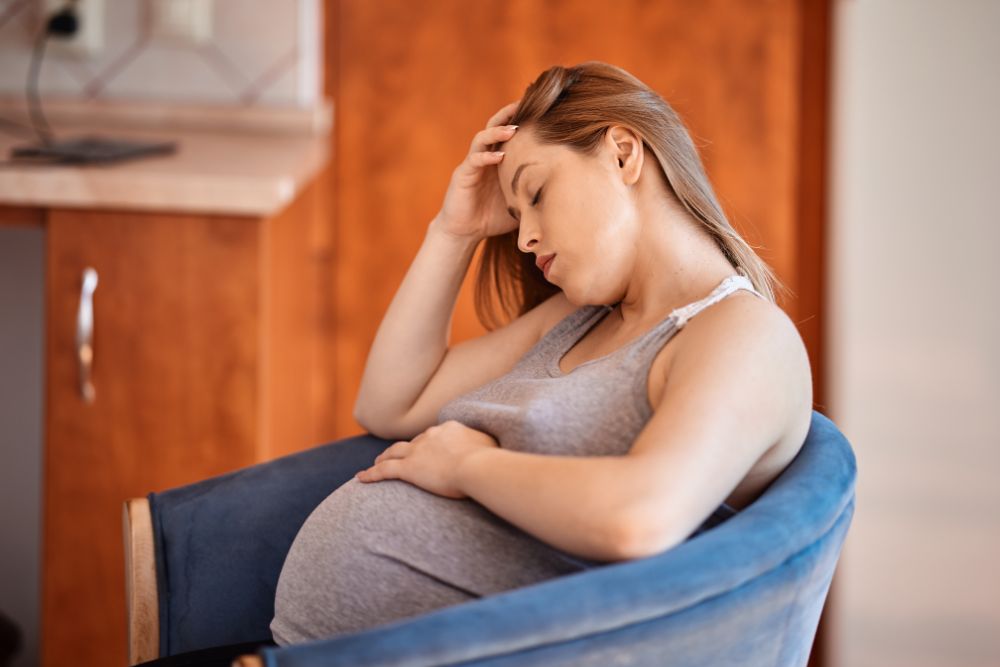 pregnant woman sitting in a chair, holding her belly and resting her head in her hand, appearing fatigued and low on energy, indicating symptoms of anemia during pregnancy.